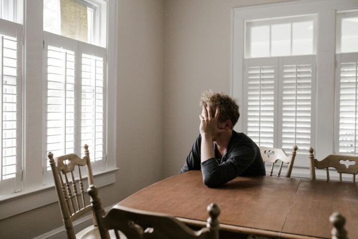 Person sitting alone at a wooden dining table with their head in their hand, appearing to experience anxiety or emotional distress in a quiet, sunlit room with white shutters