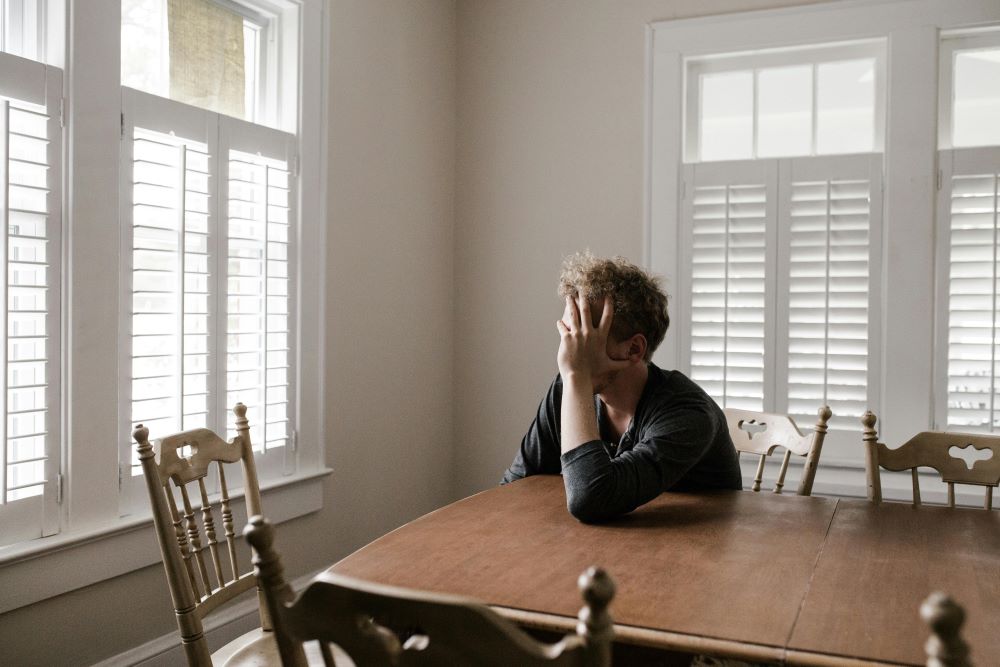 Person sitting alone at a wooden dining table with their head in their hand, appearing to experience anxiety or emotional distress in a quiet, sunlit room with white shutters