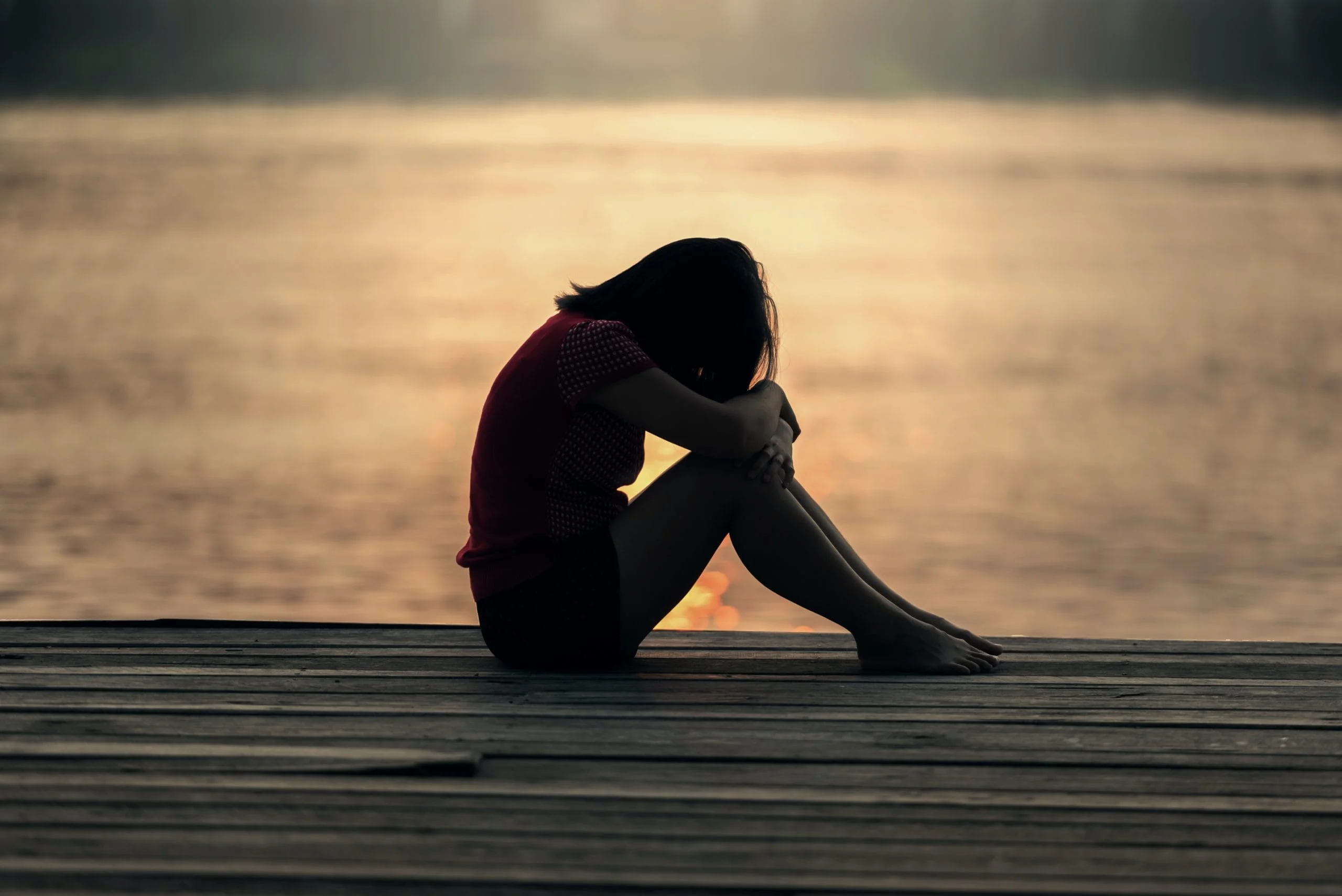 A woman sits on a dock by the water at sunset, after reflecting on her emotions during an emotional release therapy session.
