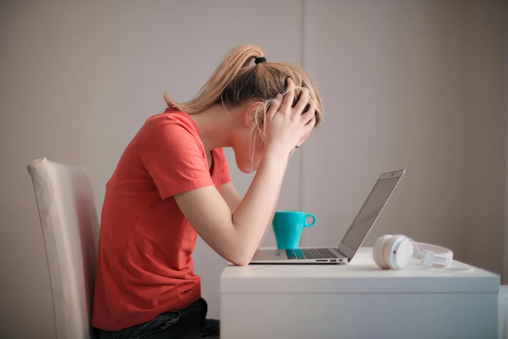 Person sitting at a desk with head in hands, appearing stressed, in front of an open laptop -struggling with overcoming perfectionism - while a blue mug and headphones rest nearby.